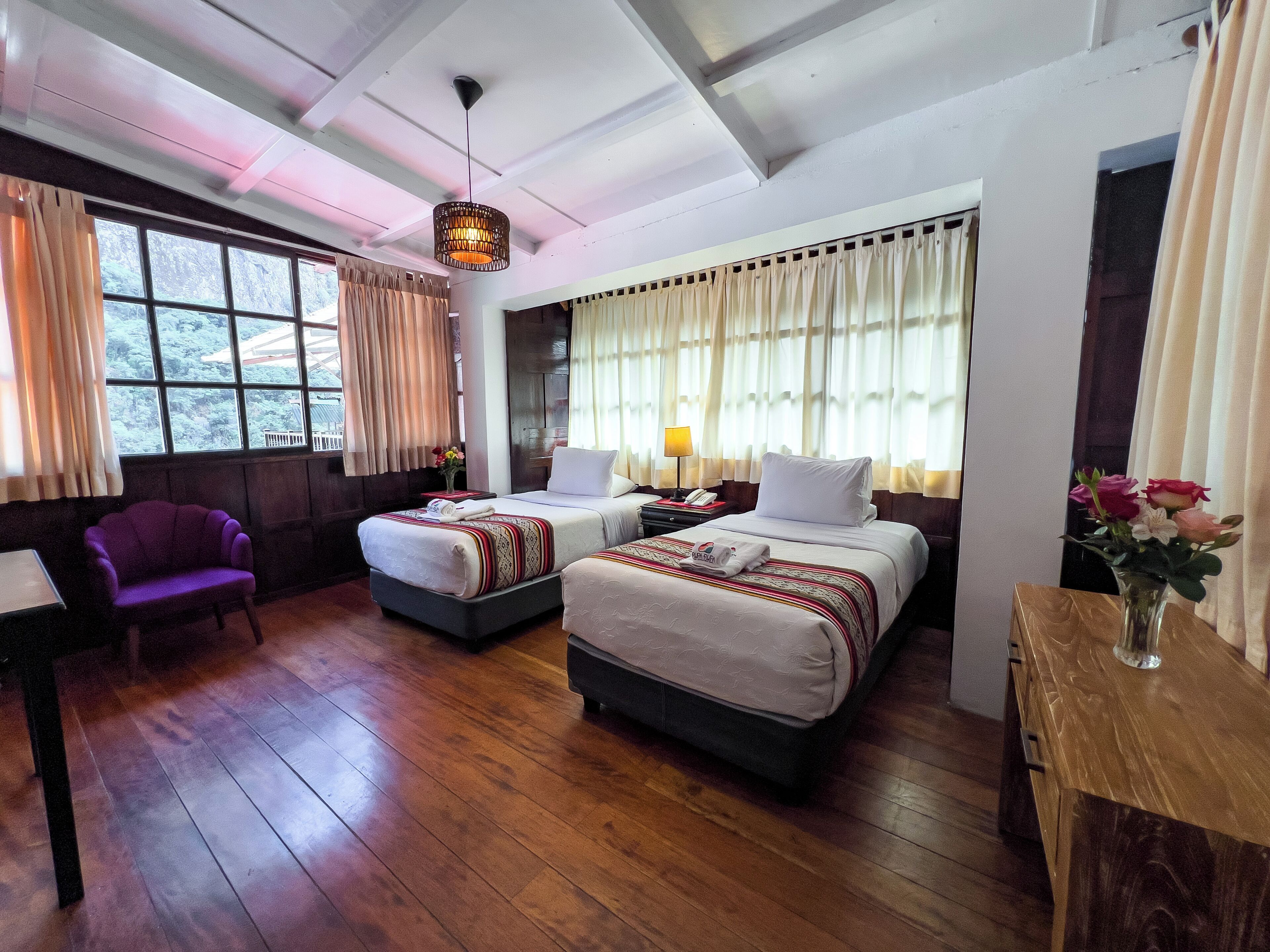 Quarto de hotel com duas camas, uma poltrona roxa, uma mesa de madeira e um vaso com flores.