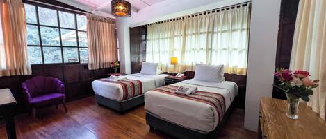 Quarto de hotel com duas camas, uma poltrona roxa, uma mesa de madeira e um vaso com flores.