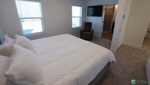 Une chambre avec un grand lit, deux fenêtres, une chaise et un téléviseur fixé au mur.