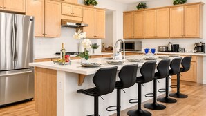 A modern kitchen with wooden cabinets, a stainless steel refrigerator, a central island with bar stools, and a microwave.