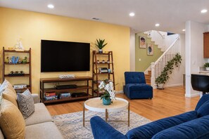 A living room with a sofa, a blue armchair, a coffee table, a TV, and shelves with books and decorative items.