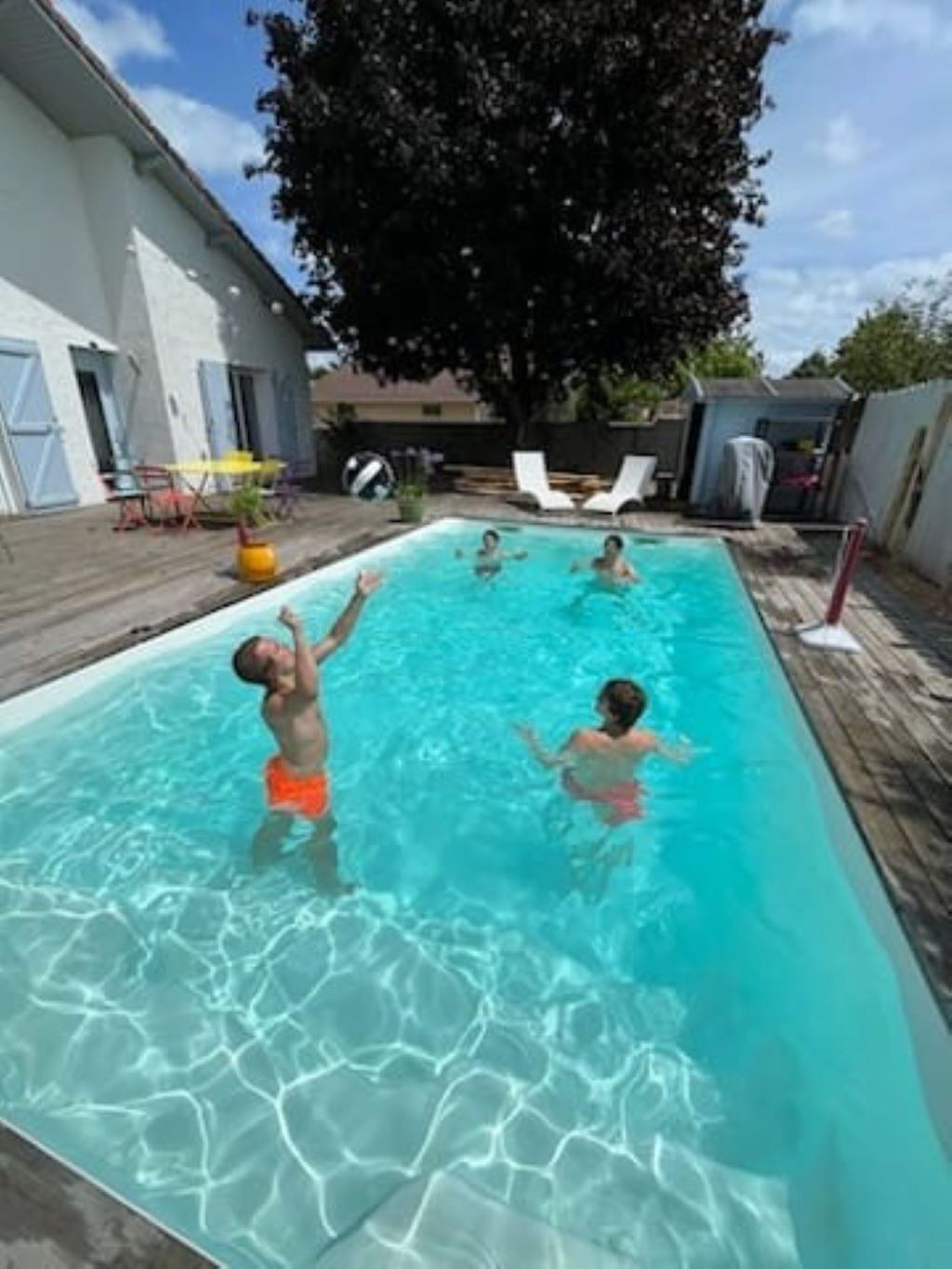 A rectangular swimming pool with clear blue water, surrounded by a wooden deck and a white building with blue shutters.