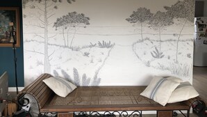 Une pièce avec une grande fresque murale représentant un paysage, un canapé en osier avec des coussins et un tableau encadré accroché au mur.