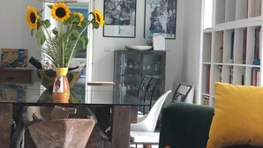 Un salon avec une table à manger en verre, un canapé vert, un coussin jaune et un luminaire suspendu en fibres tressées.