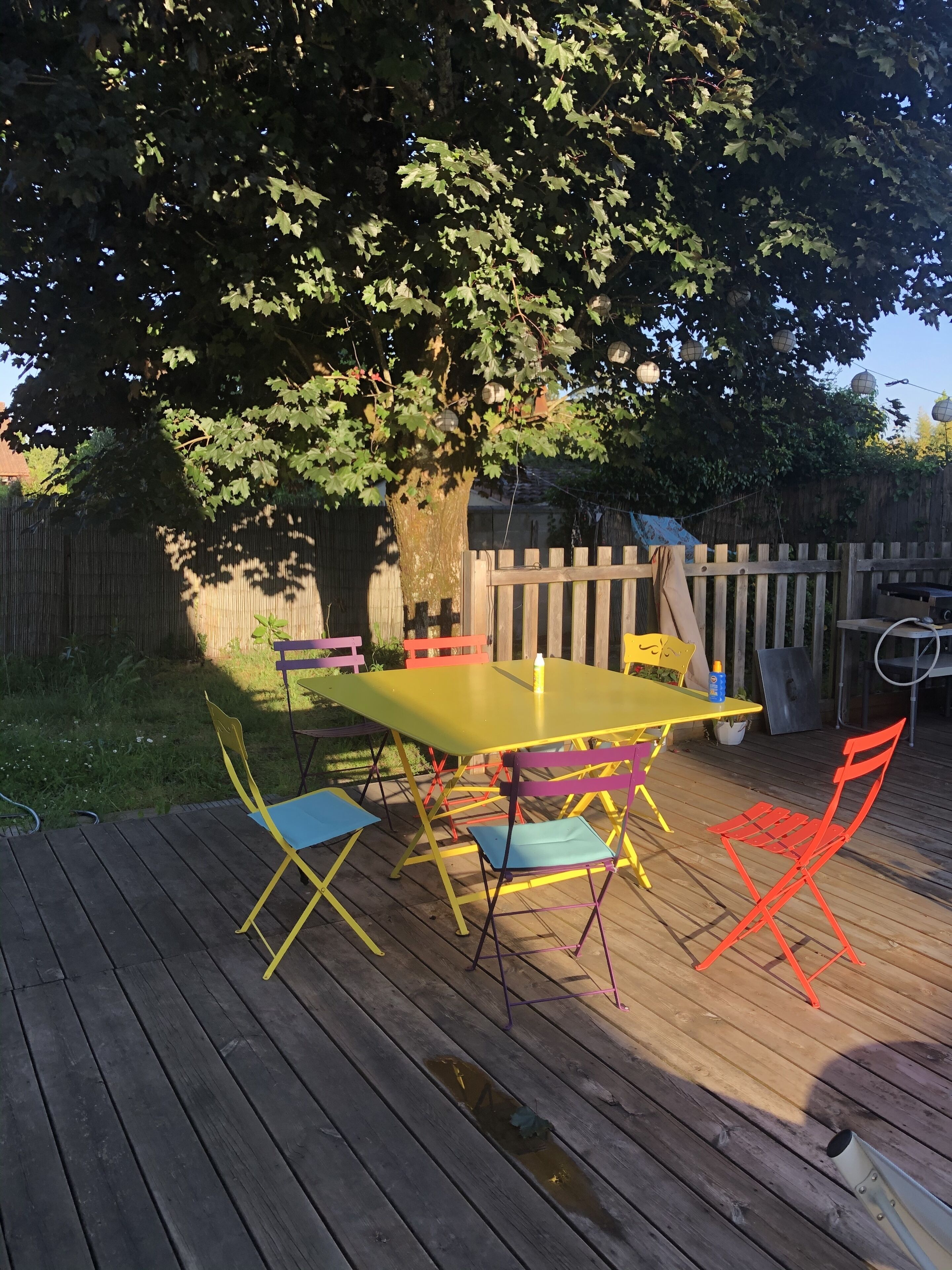 A wooden deck with a yellow table and colorful chairs, surrounded by a wooden fence and a tree.