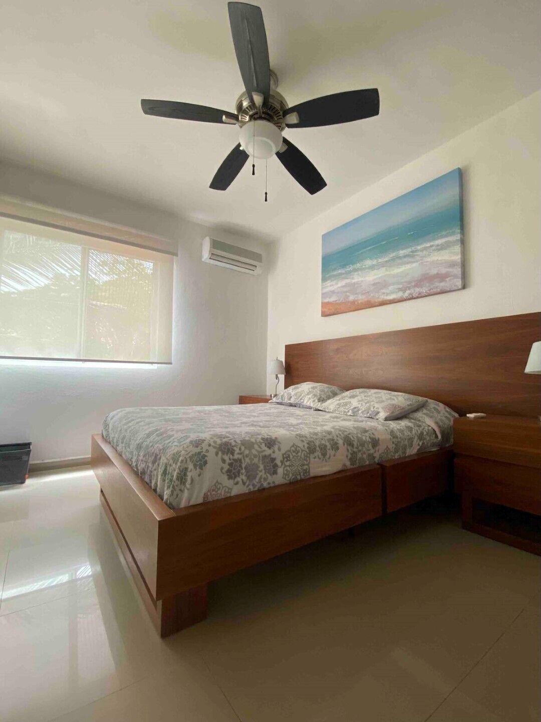 A bedroom with a bed, a ceiling fan, a painting of a beach, and a window with blinds.