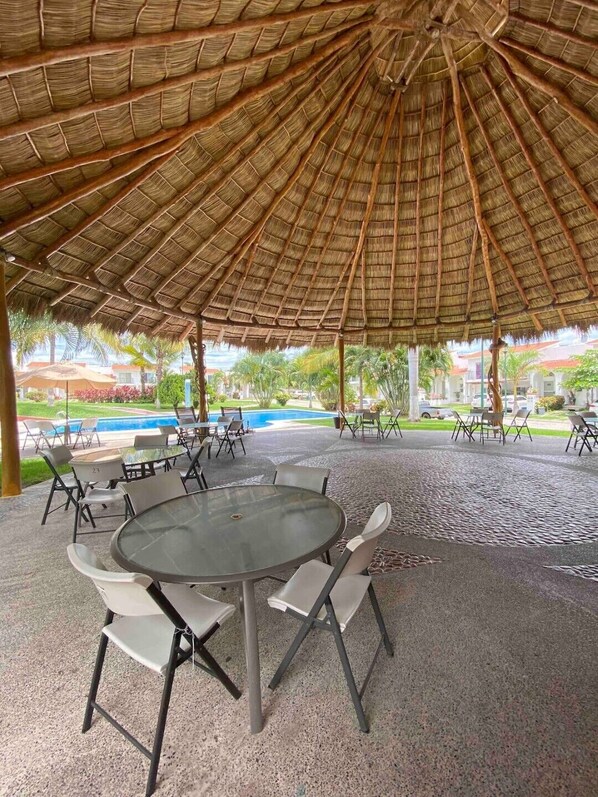 An outdoor patio with a thatched roof, round tables, and chairs.