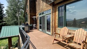 En træterrasse med udendørs siddepladser, et overdækket område og store vinduer med udsigt over det omkringliggende landskab.