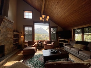A cozy living room with a stone fireplace, wooden ceiling, and large windows offering a mountain view.