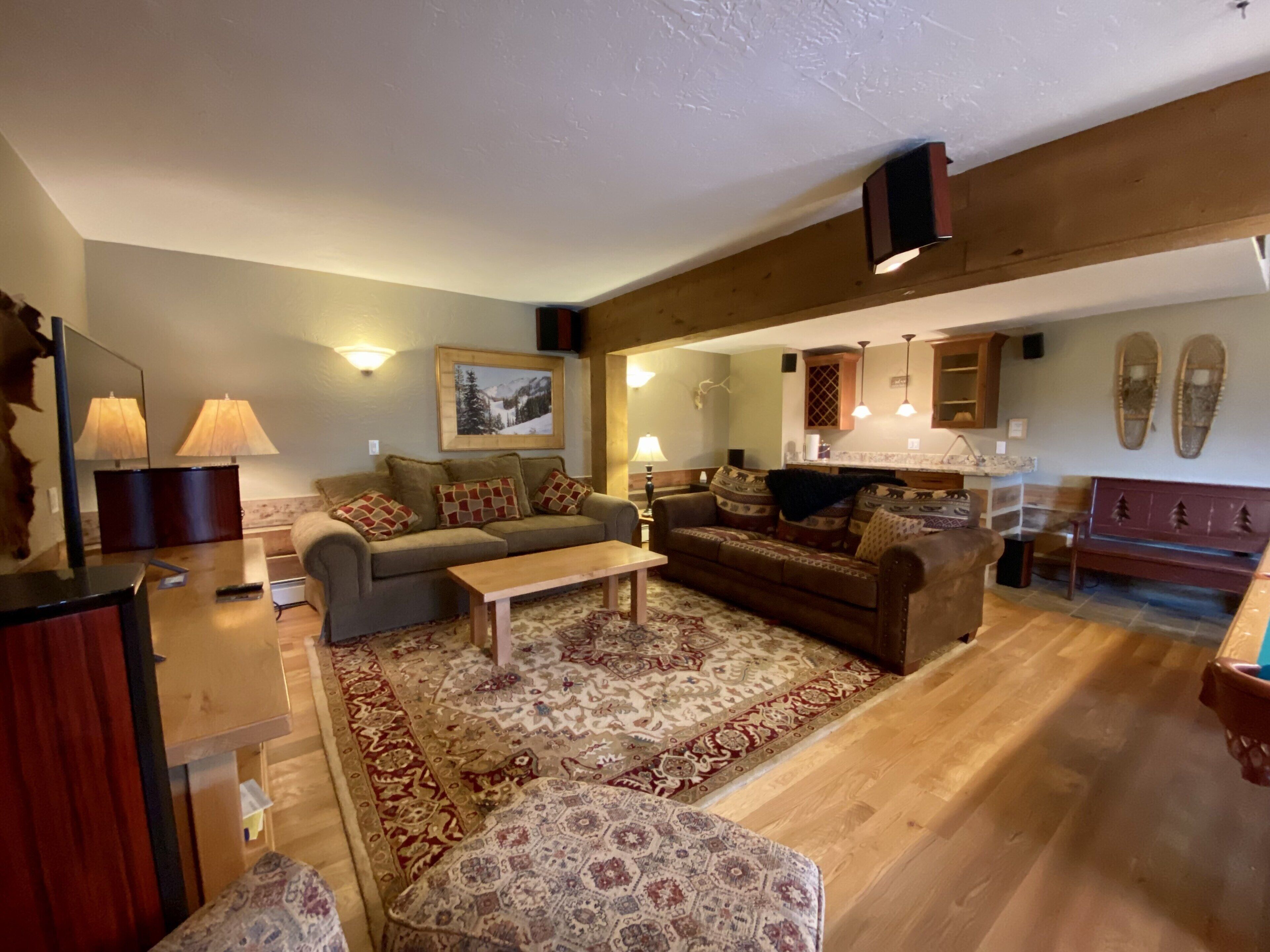 A spacious living room with a large sofa, a coffee table, and a patterned rug. There is a kitchen area with wooden cabinets and a dining table.