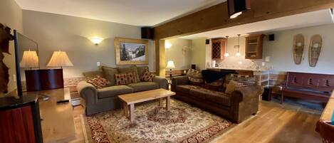 A spacious living room with a large sofa, a coffee table, and a patterned rug. There is a kitchen area with wooden cabinets and a dining table.