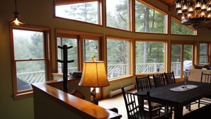 A dining area with a large table and chairs, a lamp, and multiple windows offering views of the outdoors. - Luxury Mnt. View Cabin with game room, hot tub, fire pit and private ski trails (Au Sable Forks)