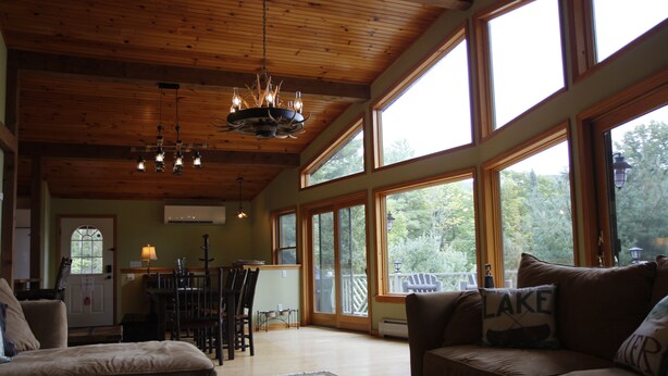 A spacious living room with a wooden ceiling, large windows, and a cozy fireplace. - Luxury Mnt. View Cabin with game room, hot tub, fire pit and private ski trails (Au Sable Forks)