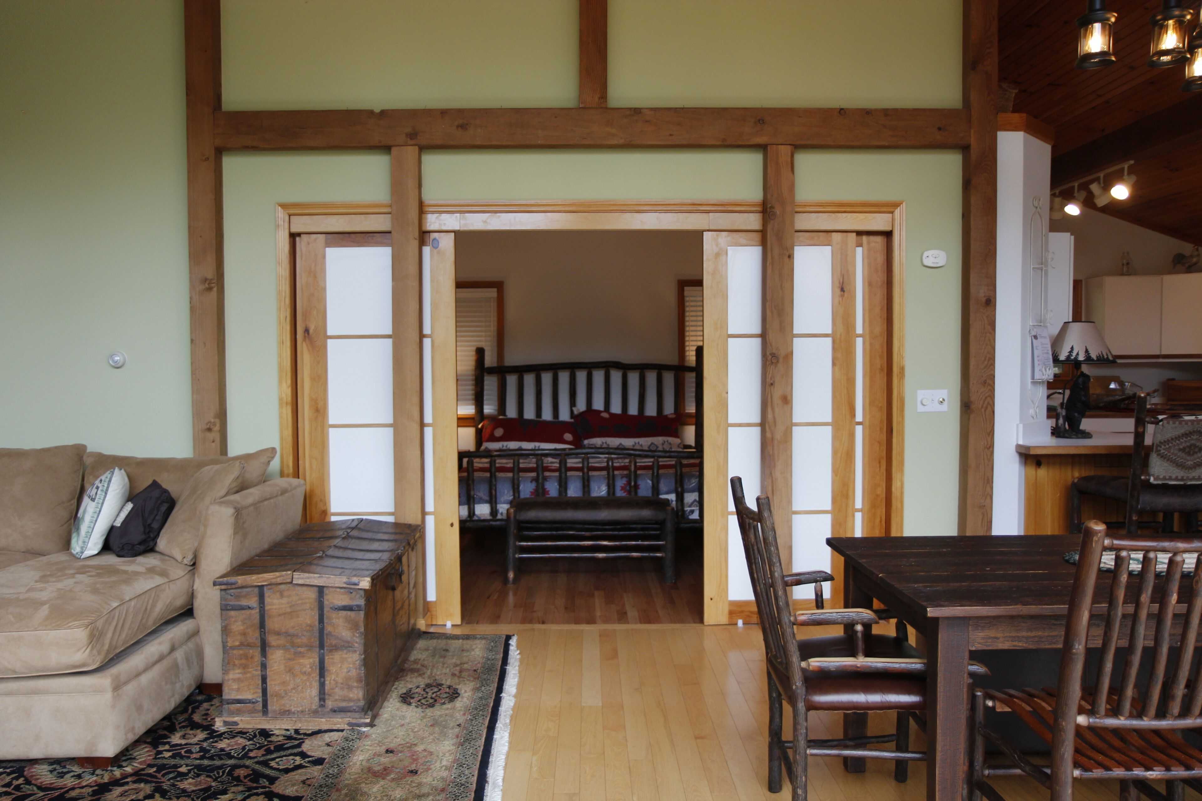 A cozy living room with a sofa, a wooden bench, and a dining table with chairs. A wooden door leads to a bedroom with a bed and a wardrobe.