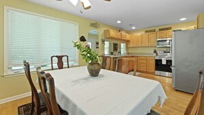 Comedor con una mesa lista para cuatro, sillas de madera, un ventilador de techo y una cocina al fondo con armarios y electrodomésticos de madera.