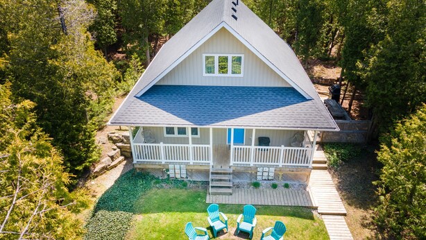 Una casa de dos pisos con fachada blanca, techo azul y una terraza. Cuatro sillas Adirondack azules están dispuestas en círculo sobre el césped delantero.
