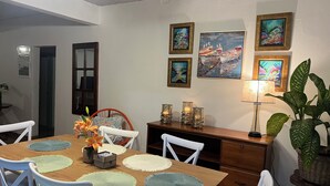 A dining area with a wooden table, white chairs, and placemats. There are framed artworks on the wall and a lamp on the side.