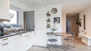 Una cocina moderna con armarios blancos, una mesa de comedor de madera y dos sillas. Hay una ventana con vistas a árboles, y una puerta que da a otra habitación.