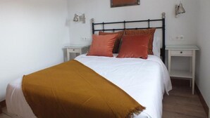 A neatly made bed with a white comforter and a mustard-colored throw blanket. There are two matching pillows and a framed picture above the bed. Two bedside tables flank the bed, each with a lamp and a small drawer. - Casa el Enebral (Colio)