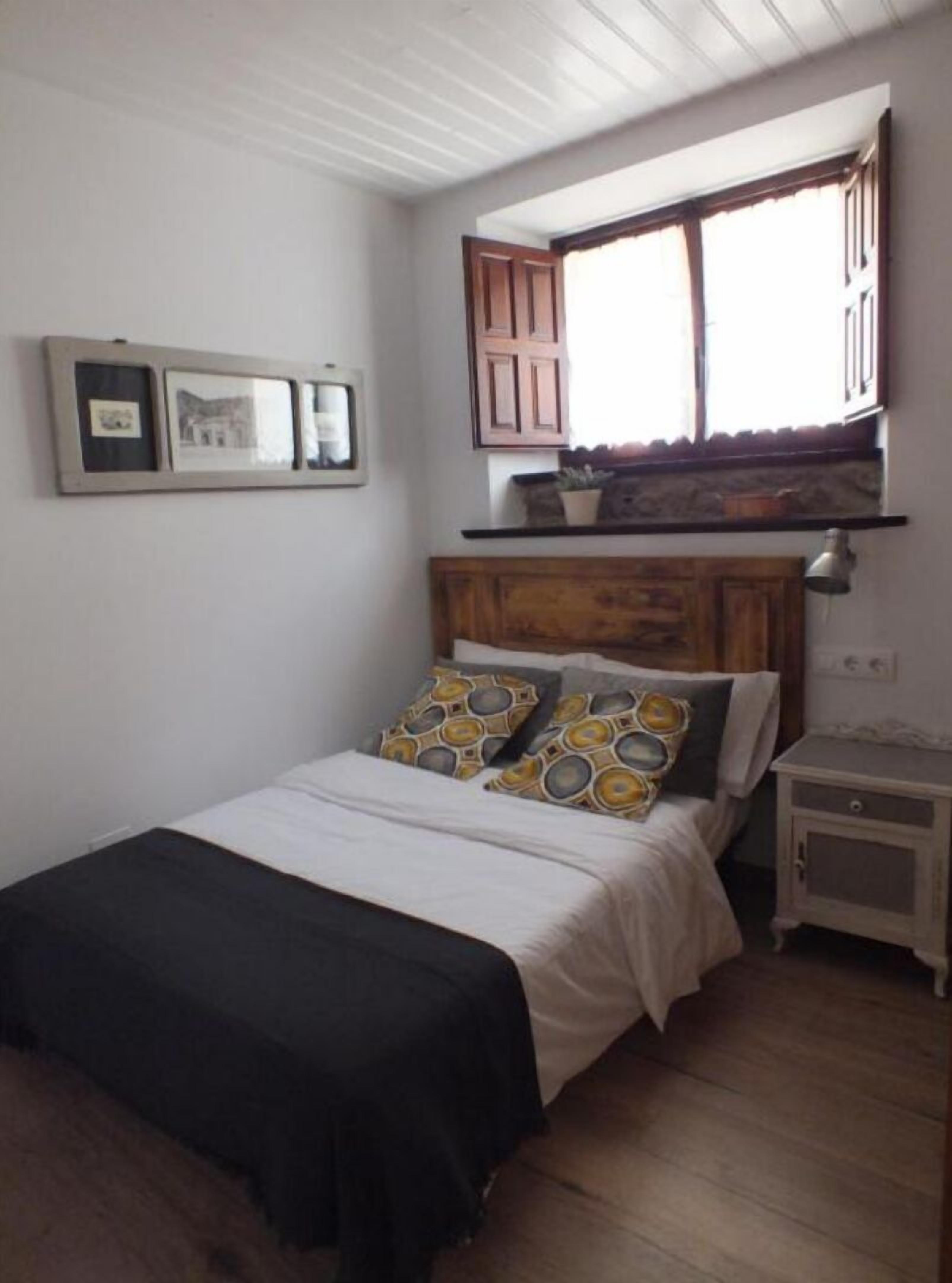 Un dormitorio con cama, mesita de noche, ventana con contraventanas de madera y repisa con planta en maceta.