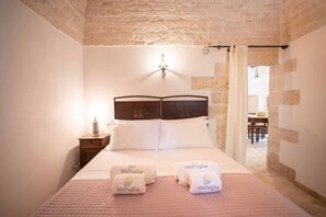 A bedroom with a stone wall, a bed with white linens, a nightstand, and a lamp.