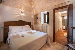A bedroom with a wooden headboard, a bed with white linens, a nightstand, a wall-mounted light fixture, and a window with a view of the hallway.