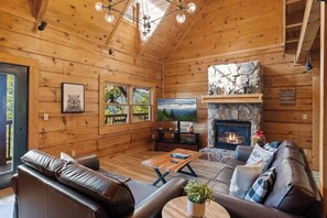 Interior de una cabaña de madera con chimenea, muebles de cuero, televisor y una pared decorativa de piedra.