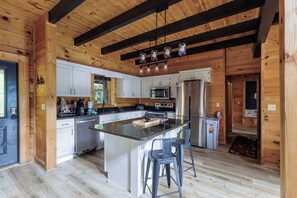 A wooden cabin interior with a kitchen featuring white cabinets, a central island, bar stools, and a hanging light fixture.