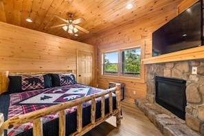Una habitación de cabaña de madera con una cama, una chimenea de piedra, un ventilador de techo y una ventana con vistas a follaje.