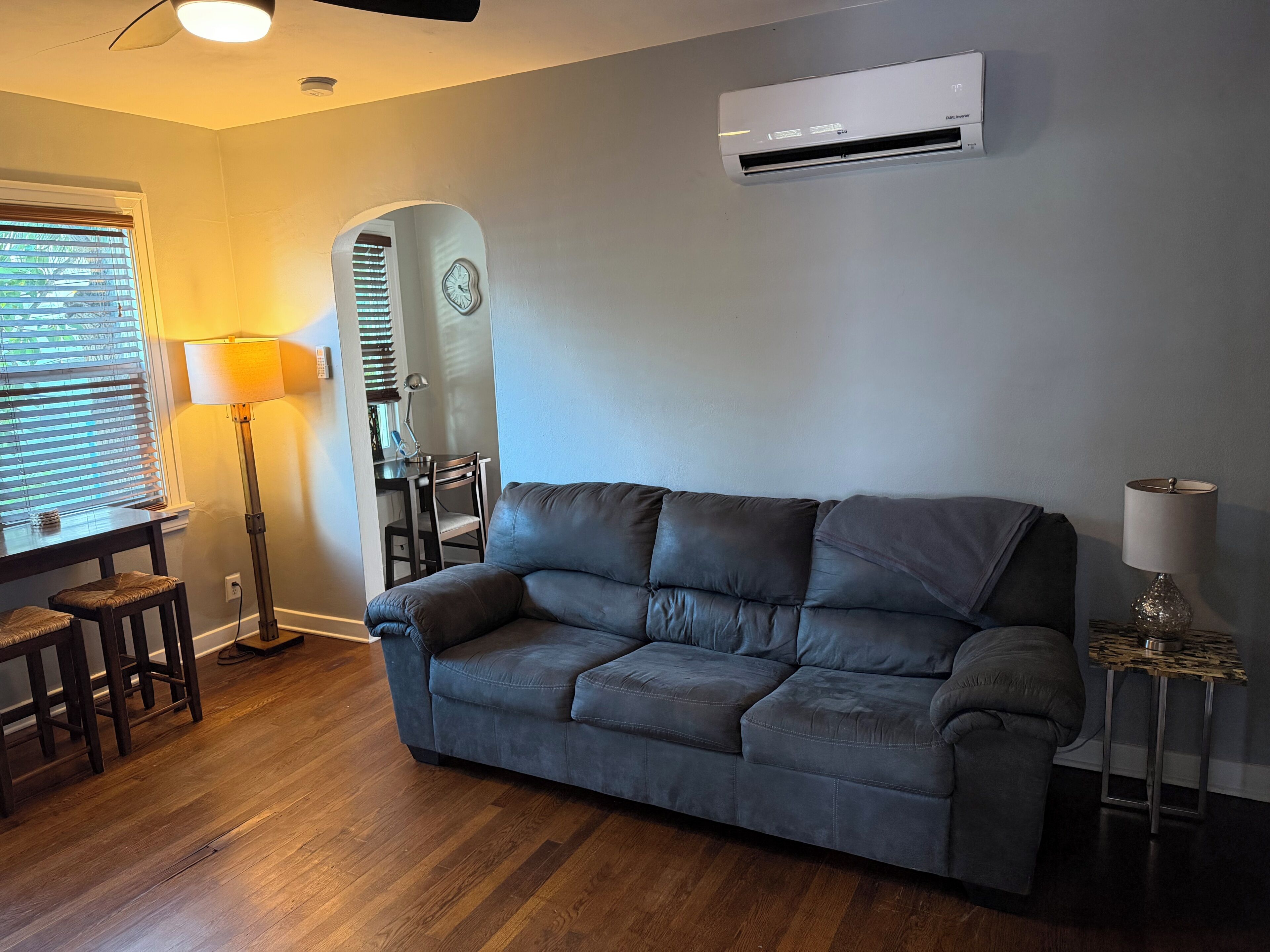 A living room with a dark grey sofa, a wooden floor, a desk with a lamp, and an air conditioning unit on the wall.