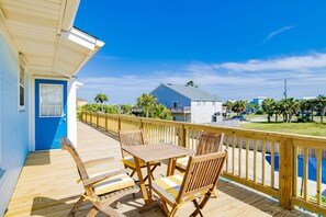 Una terraza de madera con mesa y sillas, una puerta azul y vista a una zona residencial con casas y palmeras.