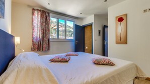 A bedroom with a bed, patterned pillows, a window with curtains, a door, and a painting on the wall. - Istron Collection Villas (Lasithi)