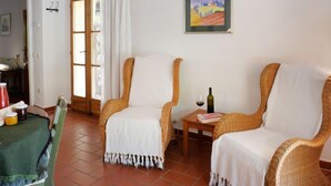 Una habitación con dos sillones de mimbre, una pequeña mesa con una botella de vino y una copa, un cuadro en la pared y una puerta que da a otra habitación.