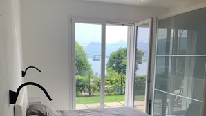 A modern bedroom with a bed, a nightstand, and a large sliding glass door leading to a balcony with a view of a lake and mountains. - Vista Unica Sulle Isole (Baveno)