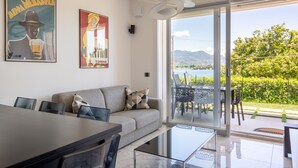 A modern living room with a sofa, a glass coffee table, and a dining area with a table and chairs. There are two posters on the wall, and a large window offering a view of greenery and a distant landscape. - Vista Unica Sulle Isole (Baveno)