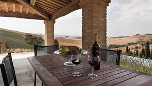 A wooden table with a bottle of wine, two wine glasses, and a bowl of fruit under a brick structure with a view of rolling hills.