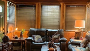 Een gezellige woonkamer met houten plafond, plafondventilator en open haard. Er zijn twee leren banken, een salontafel en een bijzettafeltje met lamp.
