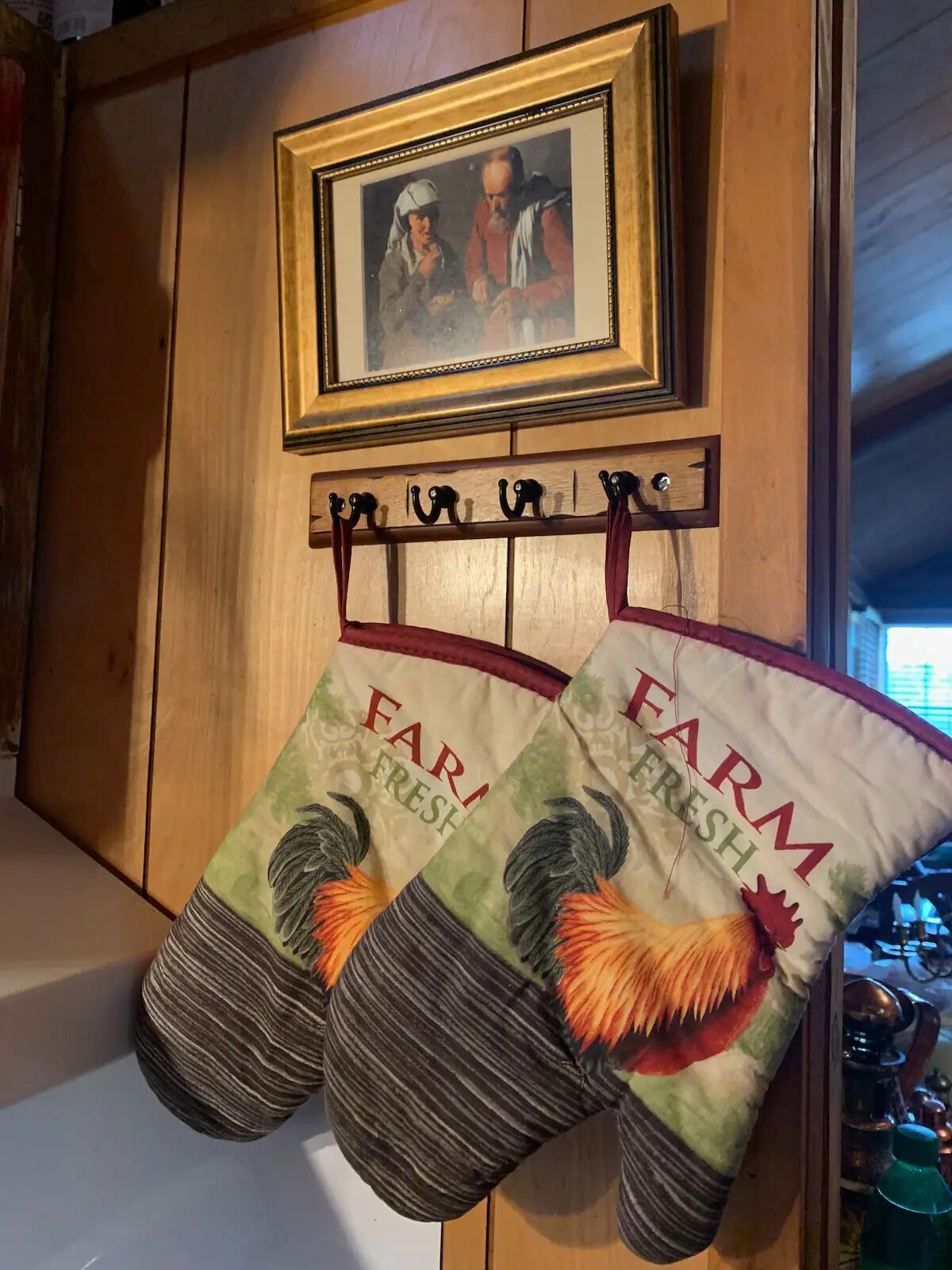 A kitchen with wooden cabinets, a framed picture above a coat rack, and two kitchen towels hanging on hooks.