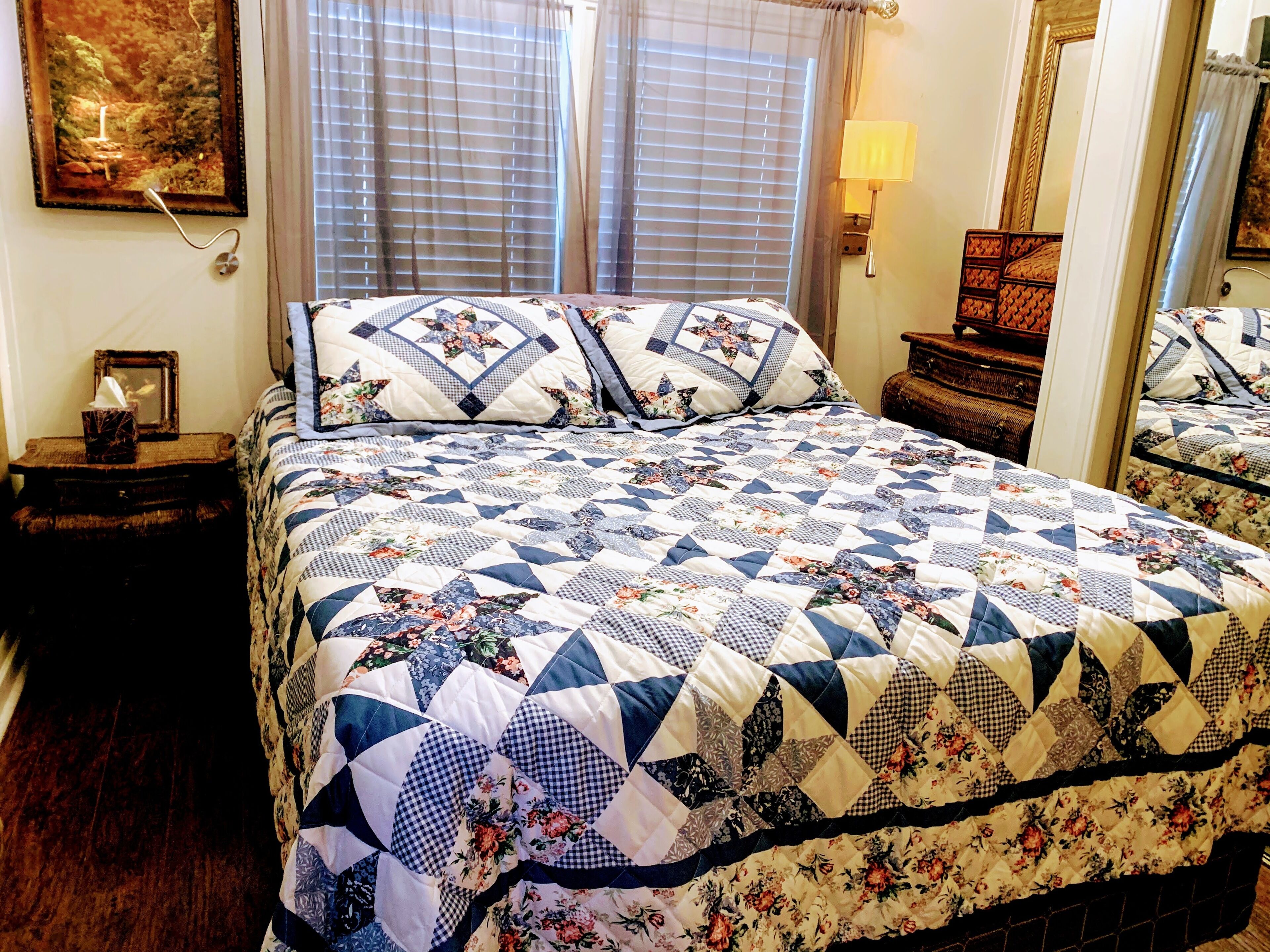 A neatly made bed with a floral and geometric patterned quilt, a bedside table with a lamp, and a framed picture on the wall.