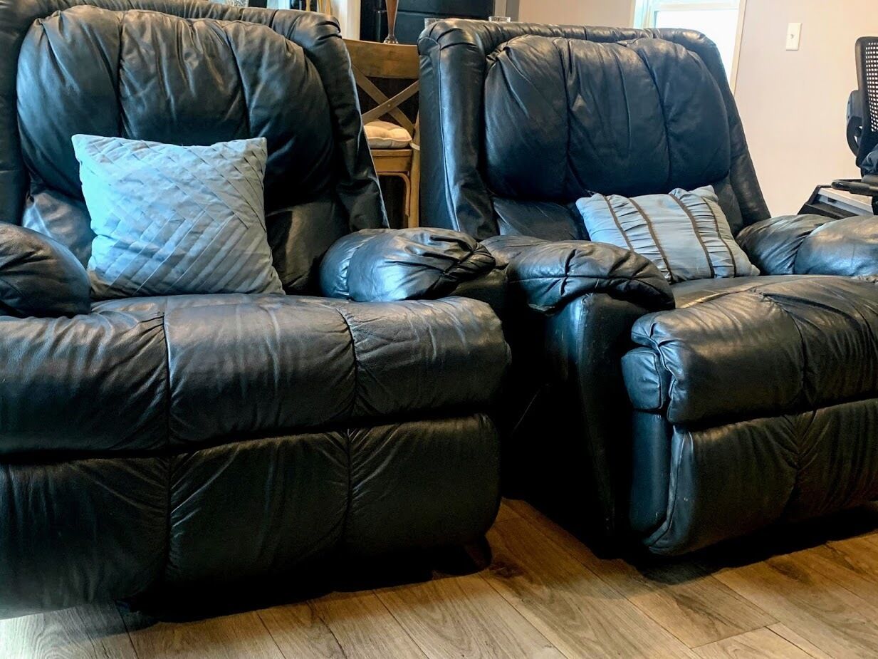 Two black leather recliner chairs with cushions, a wooden floor, and a wooden chair in the background.