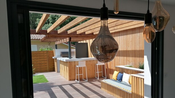 Un área de cocina exterior moderna con terraza de madera, una barra y una zona de estar con cojines.