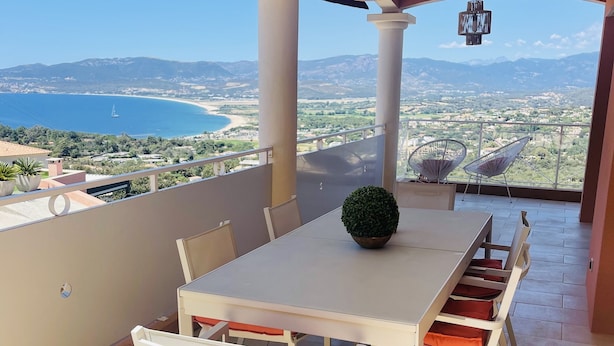 A balcony with a table and chairs, a hanging light fixture, and a view of the sea and mountains. - 3 bedroom Villa in Porticcio with Sea view (PORTICCIO)