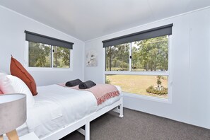 A bedroom with a bed, two windows with blinds, a painting, and a view of a garden. - Wallaby Gully Farm Cottage Peace, Quiet, Serenity (Ellalong)
