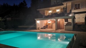 En swimmingpool med fliseterrasse, et overdækket udendørs opholdsområde med stole og et bord, samt en bygning med balkon og vinduer.
