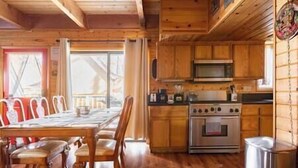 Un interior de madera con una mesa de comedor y sillas, una cocina con estufa y microondas, y un ventanal con cortinas.
