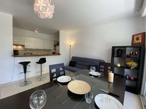 A modern living room with a glass dining table set for four, a grey sofa, and a black bookshelf with decorative items.