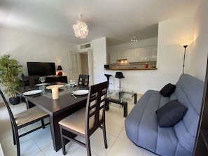 A modern living room with a grey sofa, a dining table with chairs, a TV, and a kitchen area in the background.
