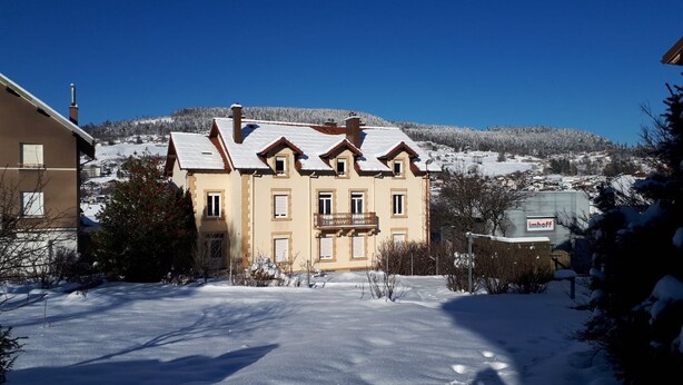 Ein zweistöckiges Gebäude mit einer symmetrischen Fassade, zahlreichen Fenstern und einem Balkon. Schnee bedeckt den Boden und die Bäume um das Gebäude, über dem sich ein klarer, blauer Himmel erstreckt.