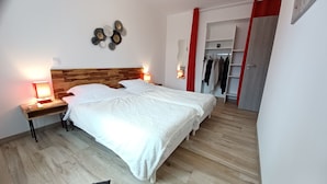 Ein Schlafzimmer mit einem Holzkopfteil, zwei Nachttischen mit Lampen, einem großen Bett mit weißer Bettwäsche, einem Kleiderschrank mit Kleidung und einem Spiegel an der Wand.