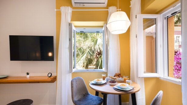 A dining area with a table set for breakfast, a TV mounted on the wall, and a window with curtains. - Corfu Town Twin Studios by CorfuEscapes (Corfu)